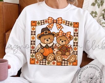 May include: A white long-sleeve sweatshirt featuring a graphic of two teddy bears inside a plaid-bordered frame. The bears wear autumn-themed clothing and bows, with one holding a pumpkin and the other a turkey. The frame is decorated with leaves.