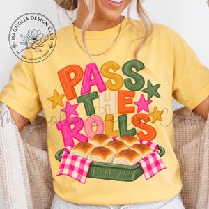 May include: A yellow t-shirt featuring the text PASS THE ROLLS in colorful, textured block letters. Below the text is a graphic of a green baking dish filled with golden-brown dinner rolls, covered by a pink and white checkered cloth napkin.