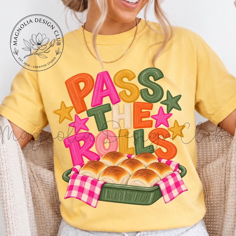 May include: A yellow t-shirt featuring the text PASS THE ROLLS in colorful, textured block letters. Below the text is a graphic of a green baking dish filled with golden-brown dinner rolls, covered by a pink and white checkered cloth napkin.