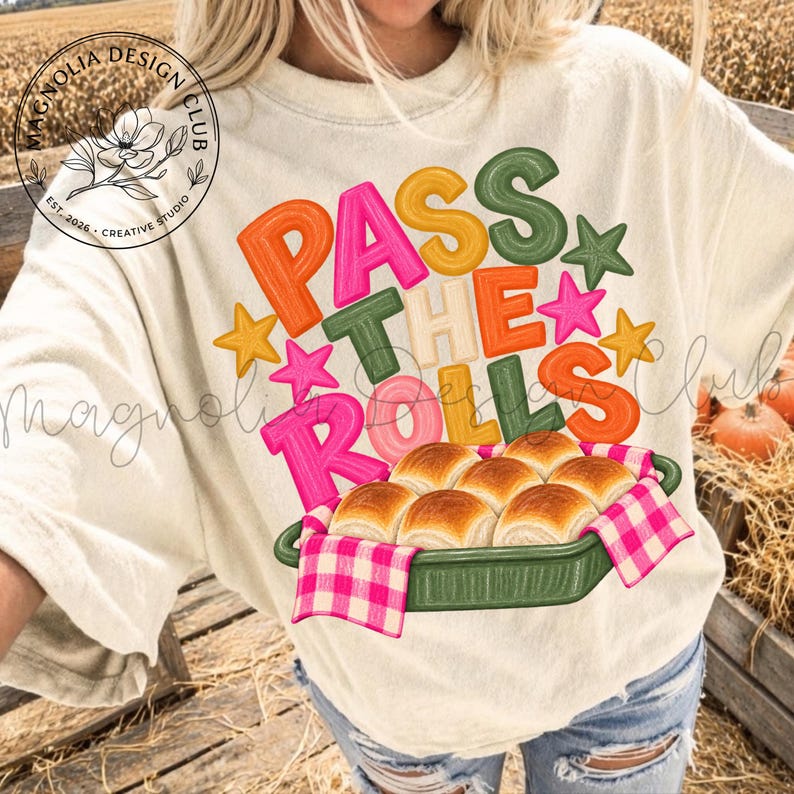 May include: A cream-colored t-shirt featuring a graphic with the text "PASS THE ROLLS" in colorful, bubble-style lettering. The design includes small stars and an illustration of golden-brown dinner rolls in a green dish with a pink checkered cloth.