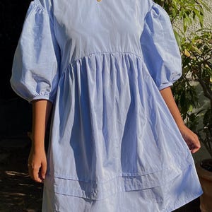 May include: A light blue babydoll dress with short, puffy sleeves. The dress features a gathered bodice and a tiered skirt. The model is wearing a red beaded necklace with a gold pendant. The dress is made of a lightweight fabric.