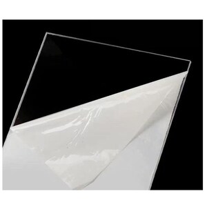 Puede incluir: Una l&aacute;mina de acr&iacute;lico transparente y clara, con una pel&iacute;cula protectora blanca parcialmente despegada. La l&aacute;mina est&aacute; inclinada sobre un fondo negro, resaltando su claridad y superficie lisa.