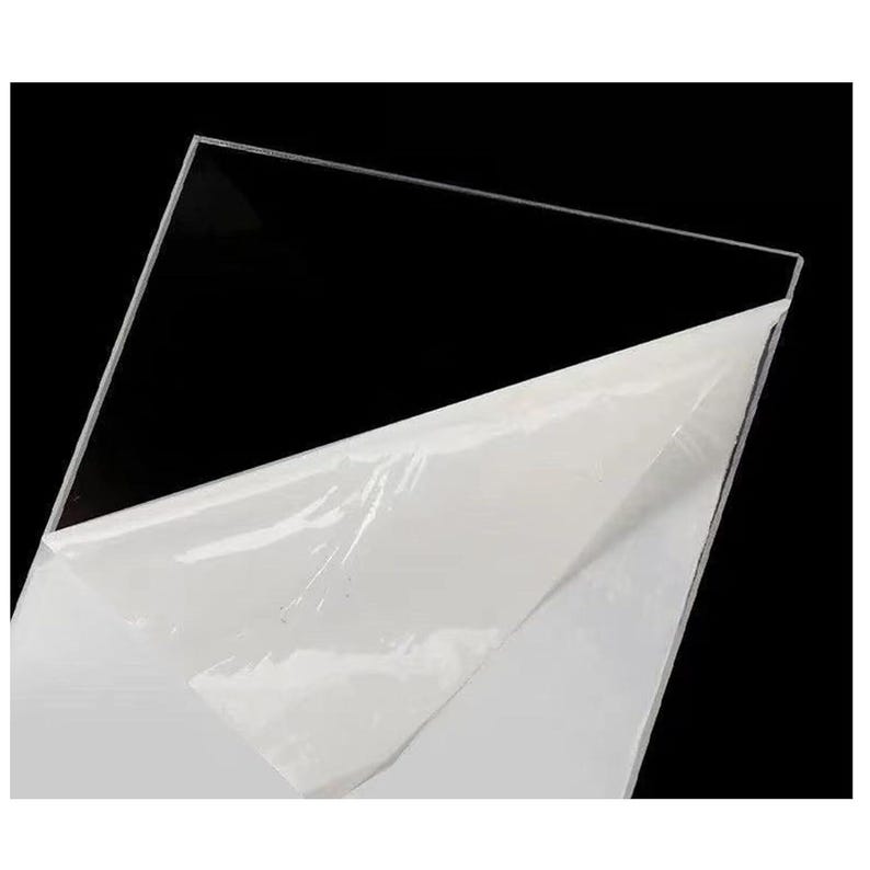 Puede incluir: Una l&aacute;mina de acr&iacute;lico transparente y clara, con una pel&iacute;cula protectora blanca parcialmente despegada. La l&aacute;mina est&aacute; inclinada sobre un fondo negro, resaltando su claridad y superficie lisa.