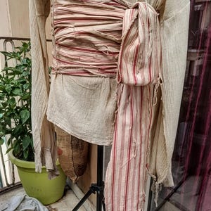 Może przedstawiać: Beżowa koszula z długim rękawem z postarzanym, warstwowym pasem owiniętym wokół talii. Pas wykonany jest z kremowego materiału w cienkie, pionowe czerwone paski, z postrzępionymi krawędziami i zwisającą częścią opadającą wzdłuż boku ubrania.