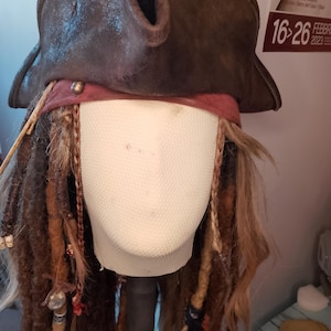 Pode incluir: Um chapéu de pirata castanho com um aspeto de couro envelhecido, aba larga e uma faixa bordô. O chapéu é combinado com longos dreadlocks castanhos adornados com contas e acessórios, criando uma fantasia.