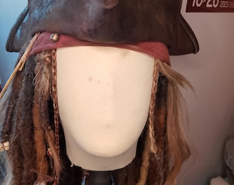 Può includere: Un cappello da pirata marrone con un aspetto invecchiato, tesa larga e una fascia bordeaux. Il cappello è abbinato a lunghi dreadlock marroni adornati con perline e accessori, creando un costume.