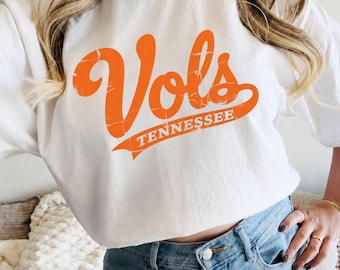 May include: A white short-sleeved t-shirt featuring the word Vols in a large, distressed orange script font across the chest, with the word TENNESSEE printed in a smaller, matching orange sans-serif font on a banner graphic underneath.