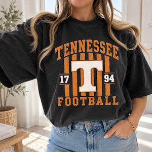 May include: A charcoal black short-sleeve t-shirt featuring a distressed orange and white graphic print. The design includes the text TENNESSEE in an arch, the number 1794, a large white letter T, and the word FOOTBALL with vertical stripes.