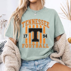 May include: A light sage green short-sleeve t-shirt featuring a distressed graphic print. The design includes the orange text TENNESSEE, a large black letter T, the numbers 17 and 94, and the orange text FOOTBALL over vertical orange stripes.