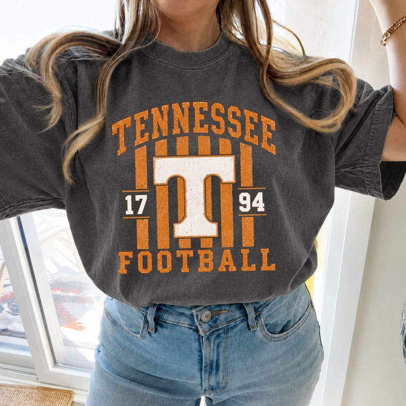 May include: A charcoal grey short-sleeve t-shirt featuring a distressed orange and white graphic print. The design includes the text TENNESSEE, the letter T, the numbers 17 and 94, and the word FOOTBALL, all arranged over vertical orange stripes.