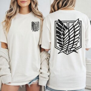 Puede incluir: Una camiseta de manga corta color blanco hueso que presenta un peque&ntilde;o gr&aacute;fico negro del emblema de las Alas de la Libertad en el pecho izquierdo y una versi&oacute;n grande del mismo emblema negro impresa en la espalda, con un dise&ntilde;o de escudo y alas.