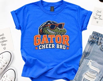May include: A bright blue short-sleeve t-shirt featuring a graphic of a green alligator wearing orange sunglasses. Below the alligator, the text reads GATOR CHEER BRO in orange and white block letters with two small orange stars.