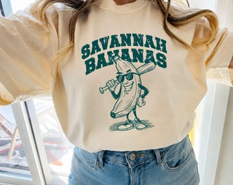 May include: A cream-colored t-shirt featuring a dark green graphic of a cartoon banana wearing sunglasses and holding a baseball bat. The text SAVANNAH BANANAS is printed in a distressed, arched collegiate font above the banana character.