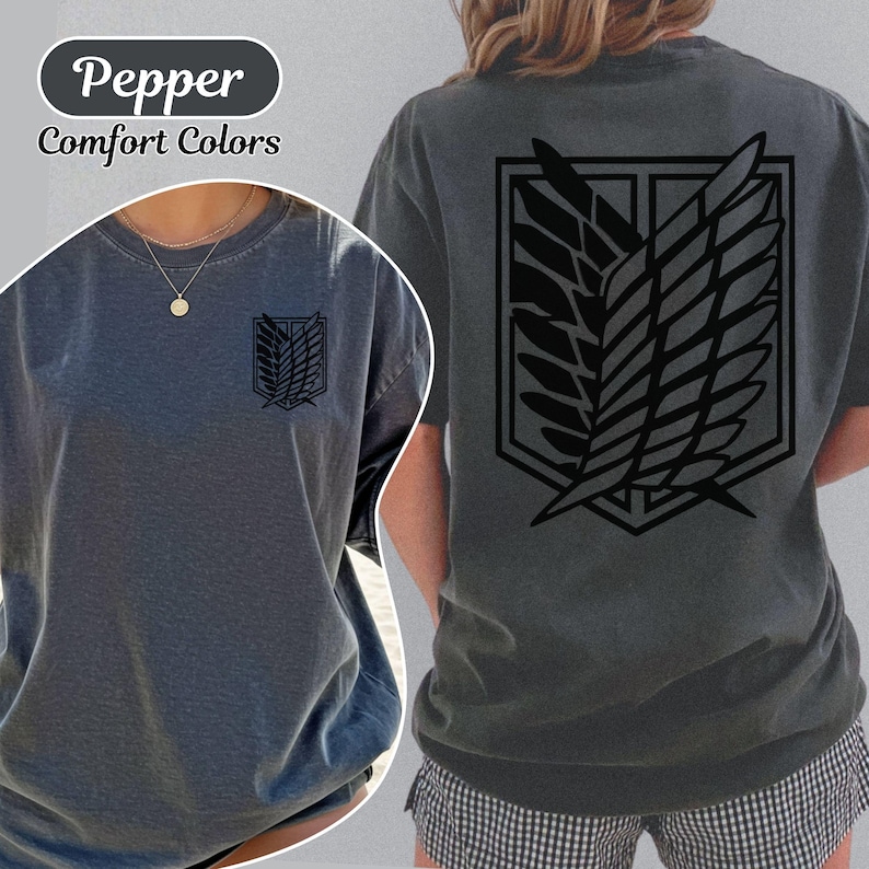 Puede incluir: Una camiseta gris carb&oacute;n con un gr&aacute;fico negro del emblema de las Alas de la Libertad en la espalda y una versi&oacute;n m&aacute;s peque&ntilde;a en el pecho izquierdo. La camiseta est&aacute; etiquetada como Pepper Comfort Colors y tiene un cuello redondo.