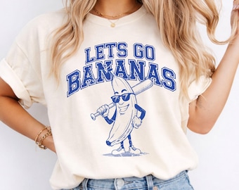 May include: A cream-colored t-shirt featuring a blue graphic of a cartoon banana wearing sunglasses and holding a baseball bat. The text above the banana reads LETS GO BANANAS in a bold, distressed blue font. The shirt has short sleeves and a crew neck.