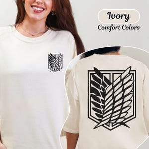 Puede incluir: Una camiseta de cuello redondo color marfil con un emblema negro de las Alas de la Libertad en el pecho izquierdo y una versi&oacute;n m&aacute;s grande del mismo emblema negro impresa en la espalda. El texto Ivory Comfort Colors es visible en la esquina superior derecha.