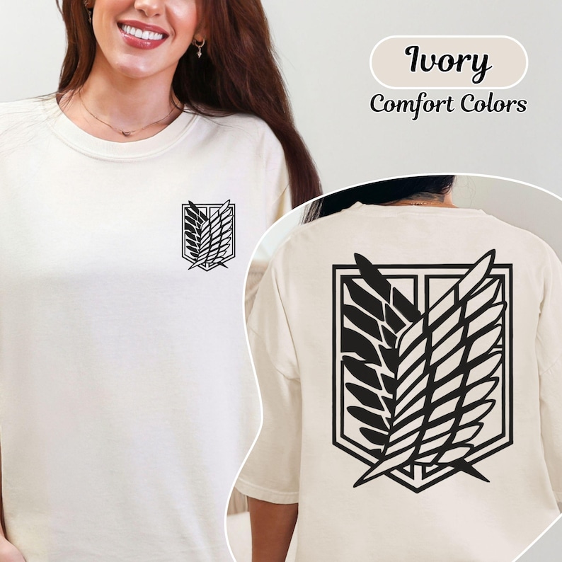Puede incluir: Una camiseta de cuello redondo color marfil con un emblema negro de las Alas de la Libertad en el pecho izquierdo y una versi&oacute;n m&aacute;s grande del mismo emblema negro impresa en la espalda. El texto Ivory Comfort Colors es visible en la esquina superior derecha.