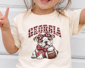 May include: A cream-colored t-shirt featuring a graphic of a bulldog wearing a red and black plaid bow on its head and a matching bow around its neck. The bulldog is holding a brown football. The word GEORGIA is printed in red and black plaid.