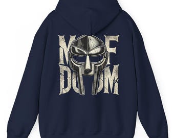 May include: A navy blue hooded sweatshirt featuring a large, distressed-style graphic on the back. The design displays a metallic-toned gladiator-style mask centered over the text "MF DOOM" in a weathered, cracked, off-white serif font.