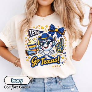 May include: Ivory-colored t-shirt with a baseball-themed graphic. The design includes a baseball character wearing sunglasses and a cowboy hat, along with the text "Go Texas!" and other baseball-related icons. The shirt is labeled "Comfort Colors."