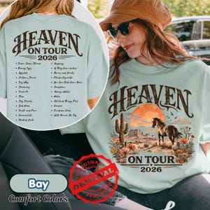 May include: A light blue t-shirt with the text "HEAVEN ON TOUR 2026" in a brown, vintage font. The front features a desert landscape with a horse, cactus, and sunset. The back lists song titles.