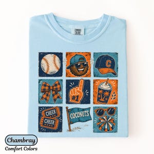 May include: A light blue t-shirt featuring a grid of nine square illustrations with baseball-themed graphics, including a baseball, a mascot, a cap, a bow, a foam finger, a drink, two tickets, a flag labeled COCONUTS, sunglasses, and a pom-pom.