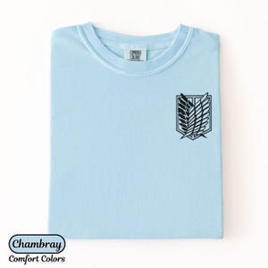 Puede incluir: Una camiseta de manga corta de color azul claro con un gr&aacute;fico negro del emblema de las Alas de la Libertad en el pecho. La etiqueta interior del cuello muestra el texto Comfort Colors. La tela es de color azul claro s&oacute;lido con cuello redondo.