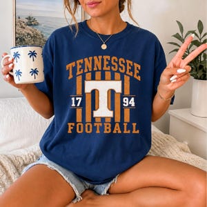 May include: A navy blue short-sleeved t-shirt featuring a graphic with the text TENNESSEE, FOOTBALL, 17, and 94, centered around a large white letter T. The design includes vertical orange stripes and a distressed texture on the lettering.