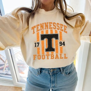 May include: A cream-colored short-sleeve t-shirt featuring a distressed graphic with the text TENNESSEE in orange, a large black letter T, the numbers 17 and 94, and the word FOOTBALL in orange, set against a background of vertical orange stripes.
