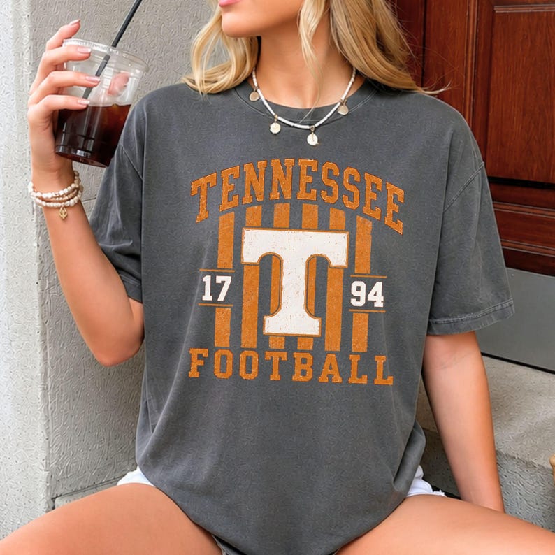 May include: A charcoal grey short-sleeve t-shirt featuring a distressed orange and white graphic print that reads TENNESSEE FOOTBALL with a large letter T in the center, flanked by the numbers 17 and 94, set against a vertical striped background.