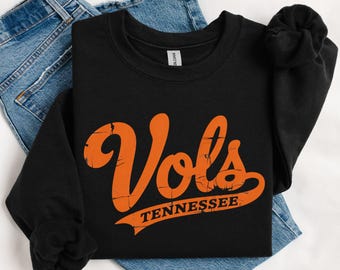 May include: A black long-sleeve crewneck sweatshirt featuring the orange text "Vols" in a large, distressed script font above the word "TENNESSEE" in a smaller, block-style font, with a curved orange banner graphic underlining the text.