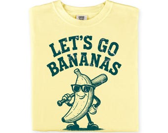 May include: A pale yellow t-shirt featuring a dark green graphic of a smiling banana wearing sunglasses and holding a baseball bat. The text above the banana reads LET'S GO BANANAS in a distressed, arched font. The shirt has a crew neckline.