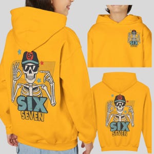 May include: Mustard yellow hoodie featuring a skeleton graphic with sunglasses and a baseball cap. The graphic includes the text "SIX SEVEN" and the number "67". The hoodie has a drawstring hood and a kangaroo pocket.