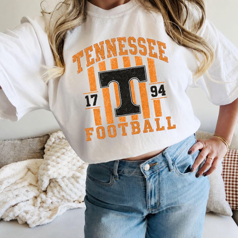 May include: A white short-sleeved t-shirt featuring a graphic design with the text TENNESSEE in orange, a large black letter T, the numbers 17 and 94 in black, and the word FOOTBALL in orange, all set against a background of vertical orange stripes.