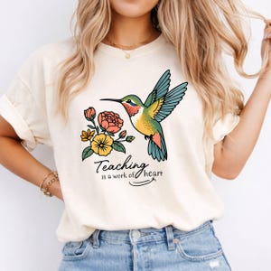 May include: A cream-colored t-shirt featuring a graphic of a hummingbird in flight next to a cluster of orange and yellow flowers. Below the illustration, the text reads, "Teaching is a work of heart" in a black, cursive, and print font combination.