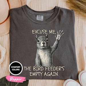 Pu&ograve; includere: T-shirt grigio scuro con grafica di uno scoiattolo che saluta, con la scritta "EXCUSE ME, THE BIRD FEEDER'S EMPTY AGAIN." La maglietta ha girocollo e maniche corte. La maglietta &egrave; di "Comfort Colors" e il colore &egrave; "Pepper".