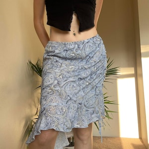 May include: A light blue paisley print skirt with a ruffled hem. The skirt has a drawstring detail on the side and a high-low hemline. The skirt is made of a lightweight fabric and has a flowing silhouette.