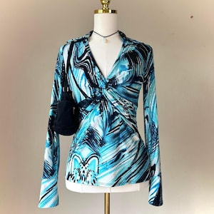 May include: A long-sleeved top featuring a vibrant blue, black, and white abstract brushstroke pattern, a V-neckline with a central twist detail, and a small black shoulder bag hanging from the mannequin's shoulder on a thin black strap.