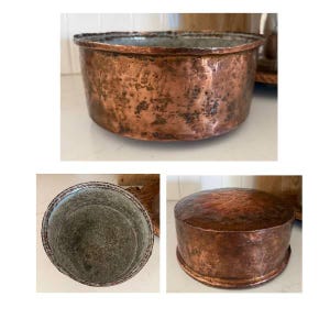 May include: A round, hammered copper container with a weathered, dark patina on the exterior and a metallic, silver-toned interior. The vessel features a matching, slightly domed copper lid that fits securely over the top of the cylindrical base.