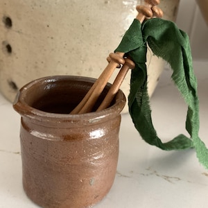 May include: A small, brown ceramic pot with a slightly irregular shape. Two wooden dowels are inside the pot, and a green fabric ribbon is tied around the dowels. The pot has a rustic, handmade appearance.