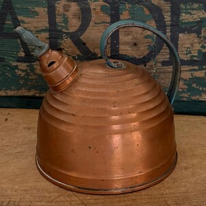 May include: A copper tea kettle with a rounded, ribbed body and a blue-toned metal handle. The spout features a decorative bird-shaped finial. The kettle has a wide base and a stepped, conical top section leading to the spout opening.