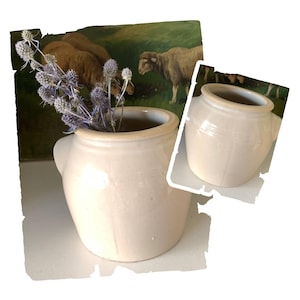 May include: A cream-colored ceramic pot with a small handle, holding dried blue flowers. A smaller version of the pot is shown to the right. The background features a painting of sheep in a field.
