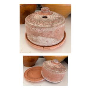 May include: A terracotta-colored, cylindrical container with a lid, sitting on a matching saucer. The lid features a decorative, etched pattern and a small knob. The container has a weathered, aged appearance, suggesting it is vintage or antique.