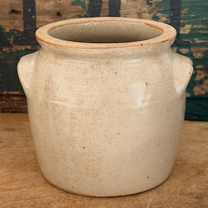 May include: A beige ceramic crock with a wide opening, a thick rounded rim, and two small integrated side handles. The exterior surface features a speckled, matte finish with subtle variations in color and texture throughout the body.