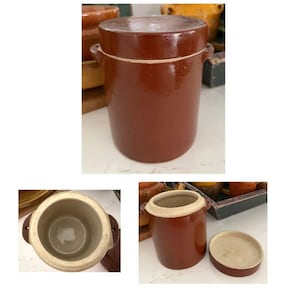 May include: A cylindrical brown ceramic crock with a matching lid and two small side handles. The exterior has a glossy dark brown glaze, while the interior and the rim are finished in a contrasting light beige, unglazed ceramic material.