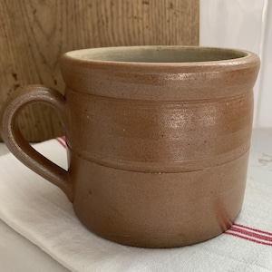 May include: A brown stoneware mug with a handle, resting on a white cloth with red stripes. The mug has a cylindrical shape with horizontal bands and a light-colored interior. The mug is likely vintage or antique.