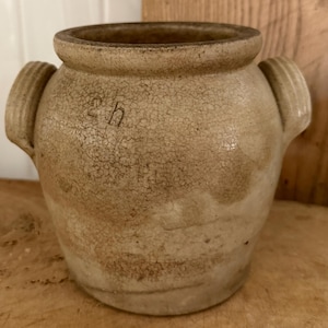 May include: A tan-colored, antique stoneware crock with two side handles. The crock has a wide opening and a slightly cracked, aged appearance. The letters "2 h" are visible on the front of the crock.