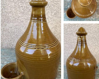 May include: A brown ceramic bird feeder with a round dish and a tall, rounded top with a knob. The feeder has a glazed finish and is decorated with brown lines.