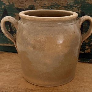 May include: A beige ceramic crock with a smooth, glazed finish and two side handles. The vessel features a wide, circular opening with a thick, rounded rim and a slightly tapered body. The surface shows subtle variations in color and texture.
