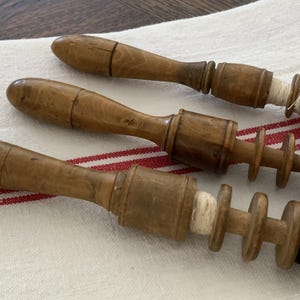 May include: Three antique wooden thread winders with a spool design, resting on a white cloth with red stripes. The winders are made of light brown wood and have a handle and a spool section. One winder has white thread, another has brown thread, and the last has black thread.